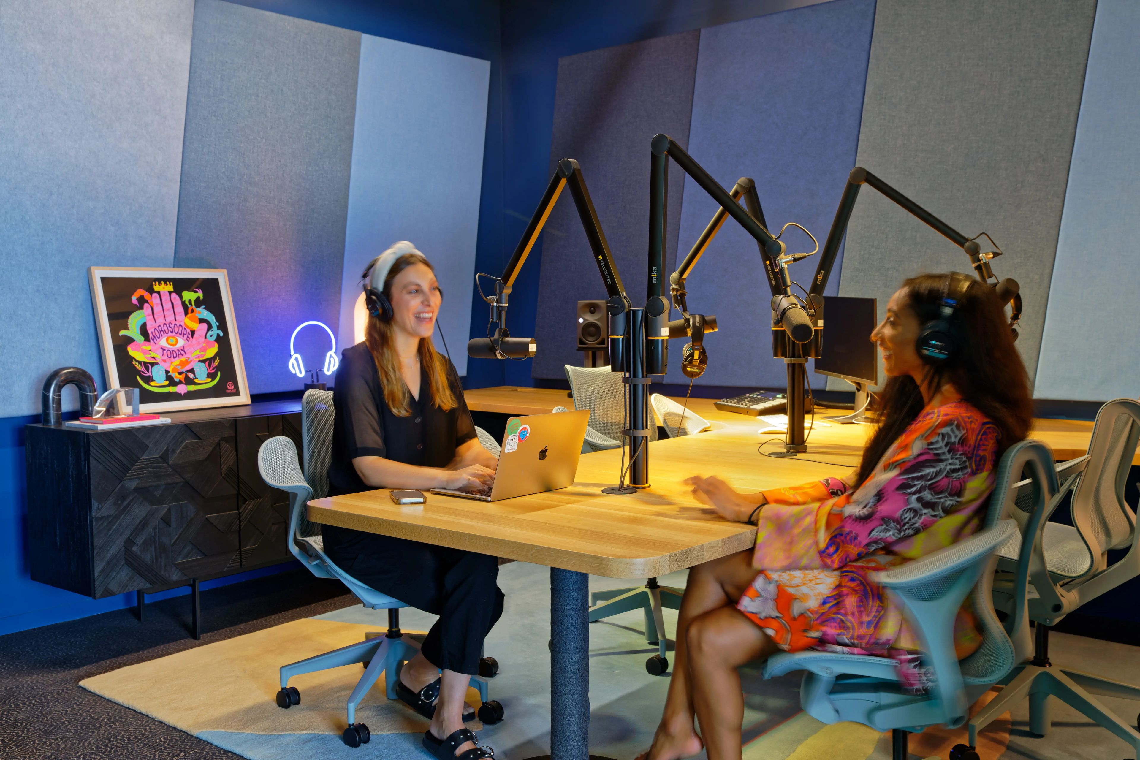 A podcast recording booth with two women seen chatting with headphones on