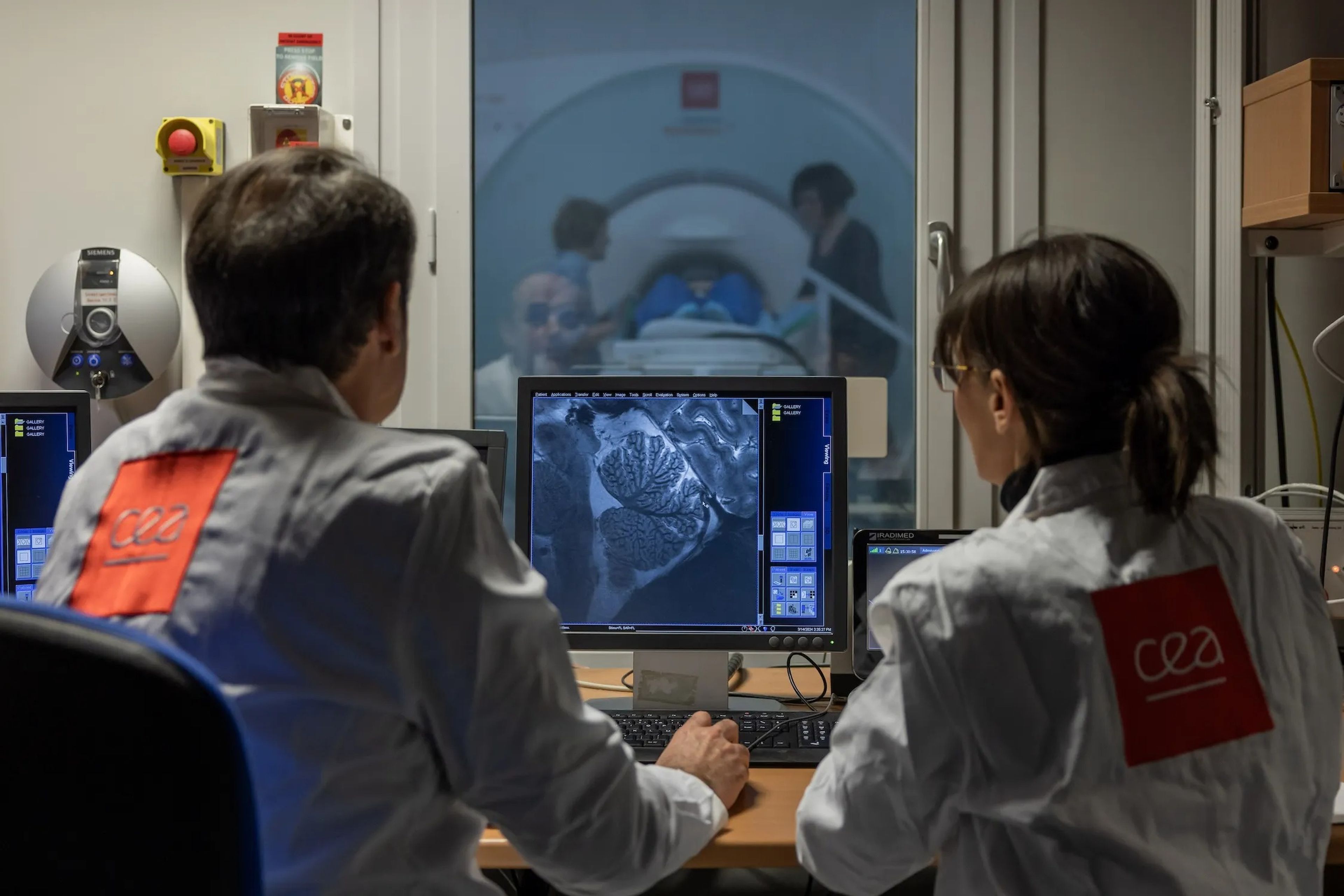 Two people sit at desk looking at a brain scan on a computer from a powerful MRI machine
