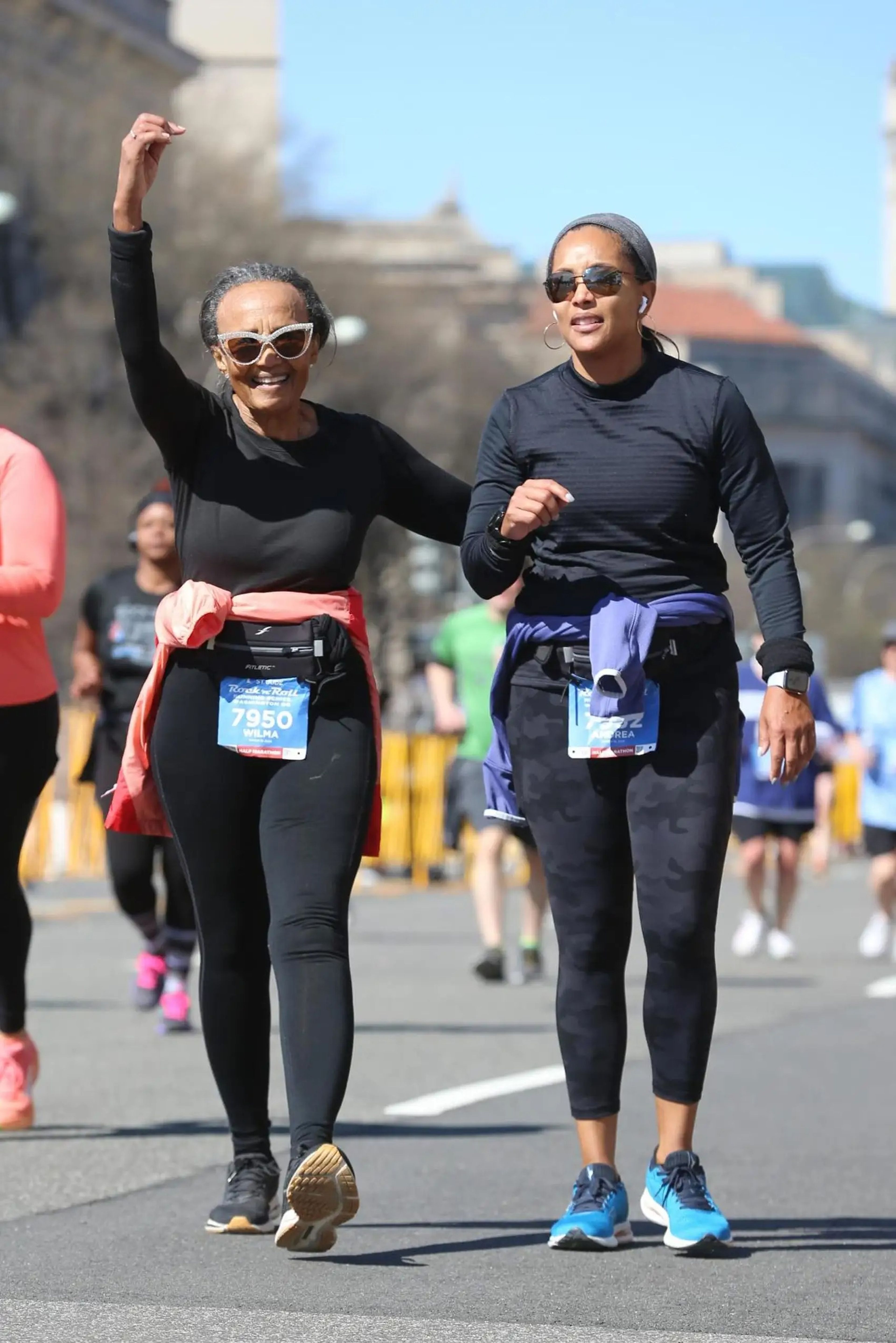 Two women in workout clothes, sunglasses, and race bibs on the road during a half marathon