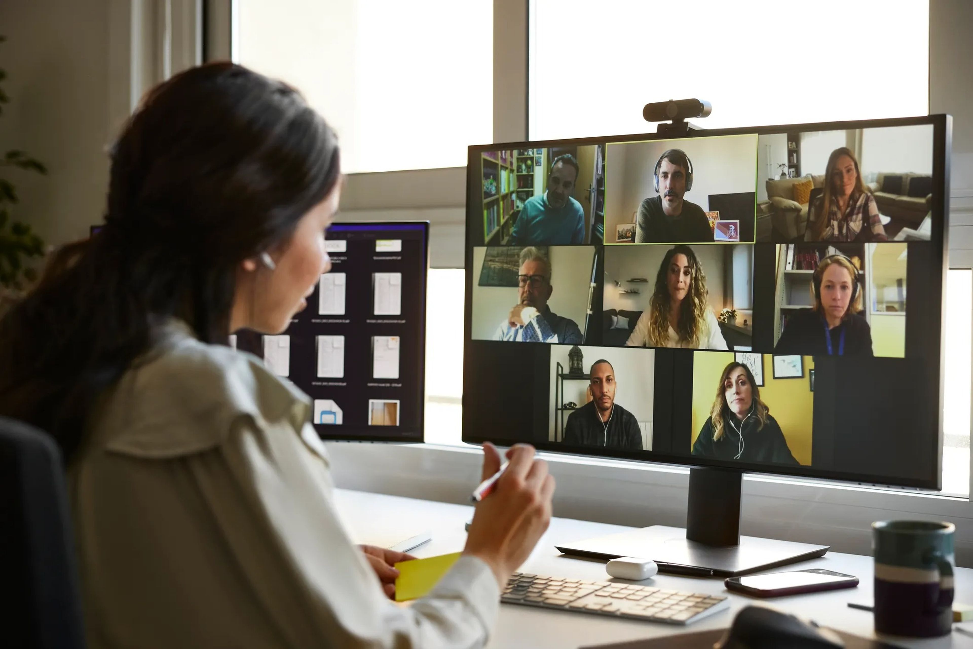 A woman at her desk participating in a video call with multiple other participants visible on the screen.