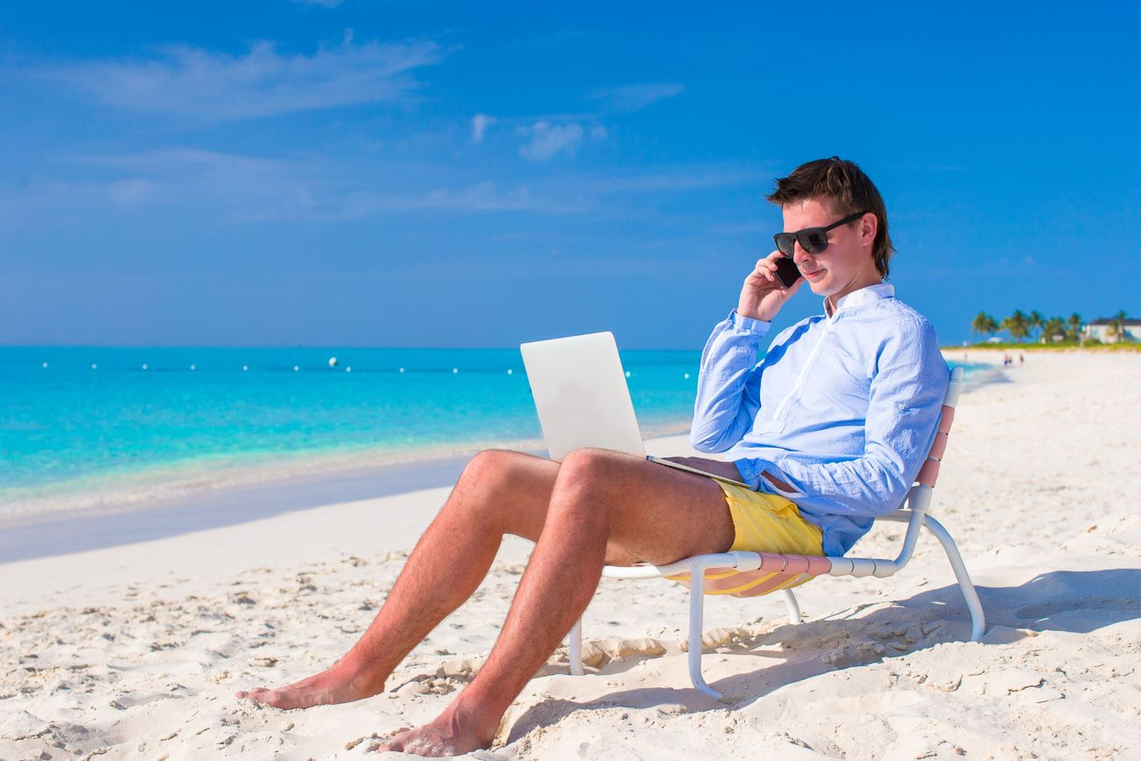 Playa, hombre joven trabajando en vacaciones con ordenador y móvil