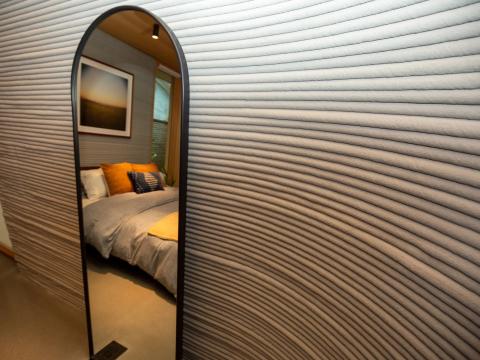 A mirror leaning against a 3D printed wall reflecting a bed.