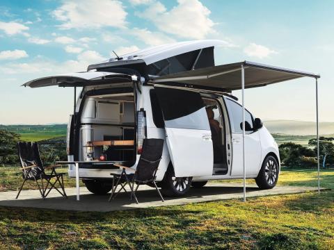 The Hyundai Staria Lounge Camper parked outside with the pop topped and awning extended. There's a table and chairs in the rear.
