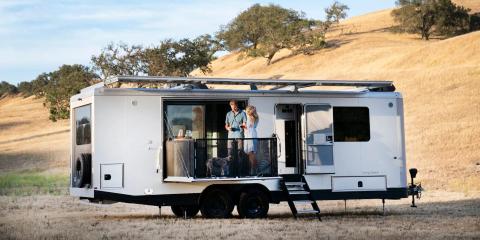 The exterior of the travel trailer as it sits on a brown field. The patio is extended and people are standing on it.
