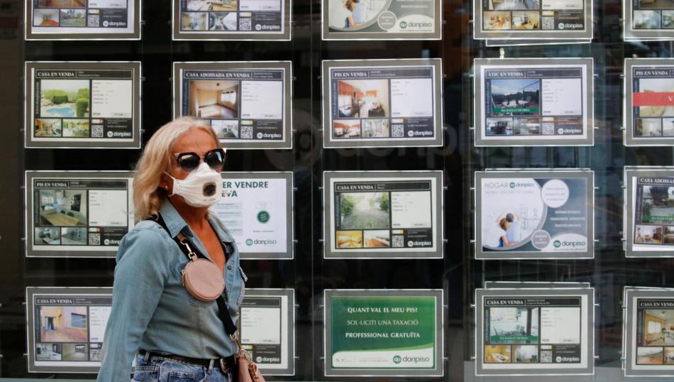 Una mujer con mascarilla pasa ante la fachada de una inmobiliaria con anuncios de pisos en venta y alquiler